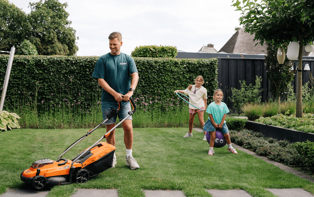 Man duwt een oranje grasmaaier over een net gazon terwijl twee meisjes achter hem spelen: één zit op een springbal, de ander zwaait met een hoepel in een besloten tuin.